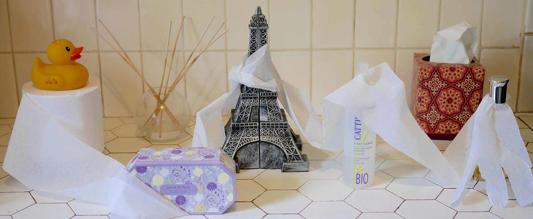 Roll of thin bathroom tissue topped by a yellow rubber ducky and draped around French soap, an Eiffel tower, and French facial spray and perfume