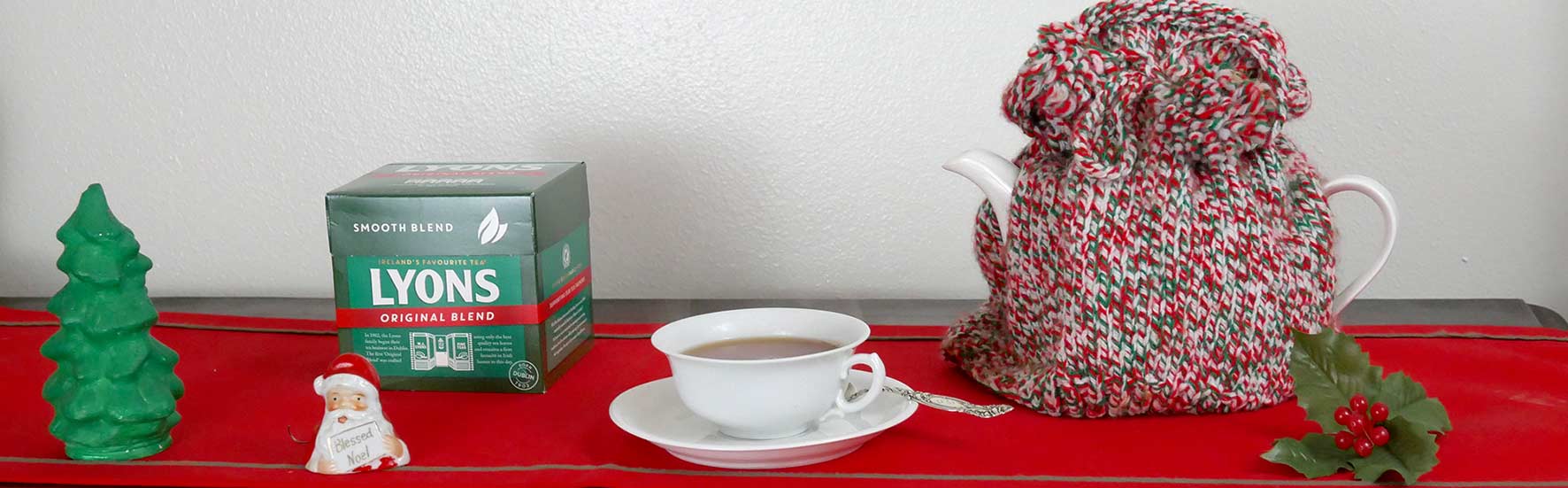 Lyons tea box and cup of brewed tea on red table runner with pot of tea covered with red, white and green tea cozy